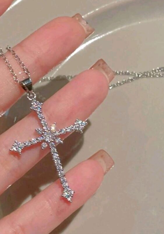 Silver cross pendant with rhinestone embellishments held by fingers, showcasing intricate design and sparkling stones on a delicate chain.